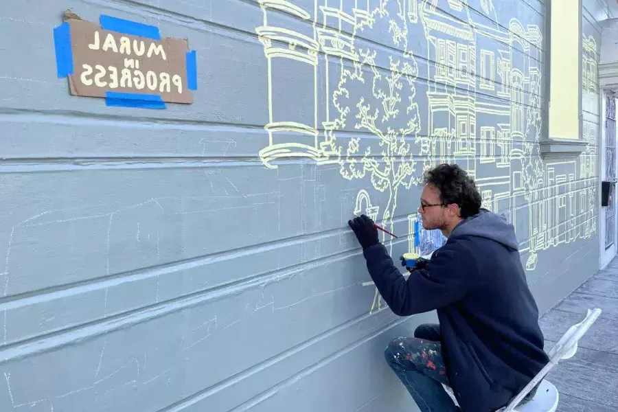 An artist paints a mural on the side of a building in the 任务地区, with a sign taped onto the building that reads "Mural in Progress.“贝博体彩app，CA。.